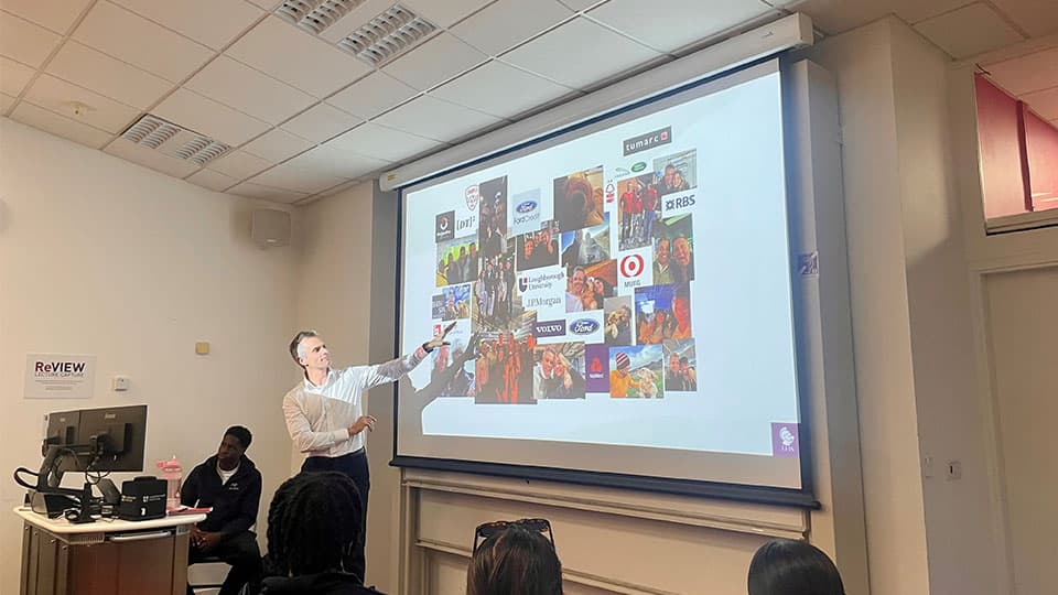 Alumnus Paul standing in front of students talking and pointing at a screen with a presentation on it.